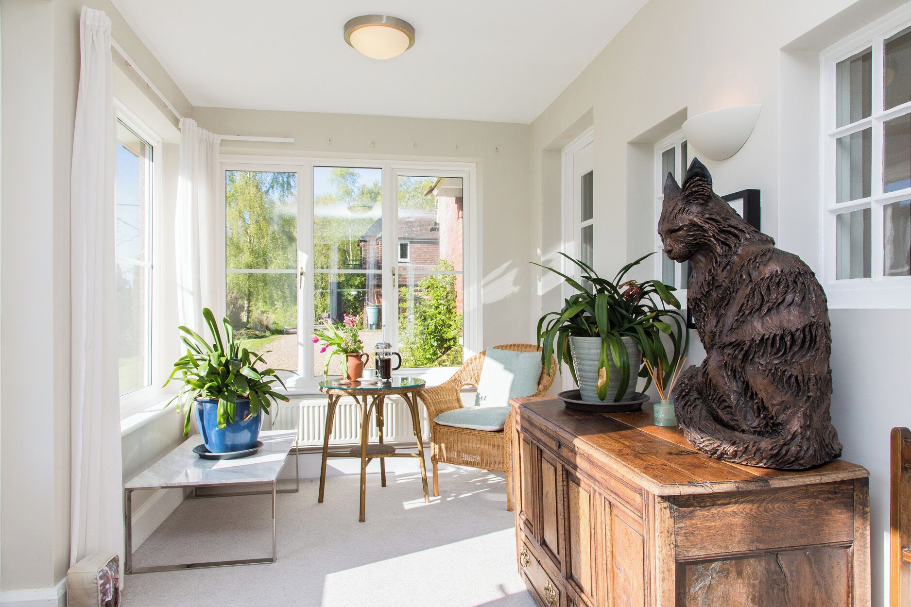 A sunlit room with large windows, a wicker chair, a wooden cabinet, and a cat statue.