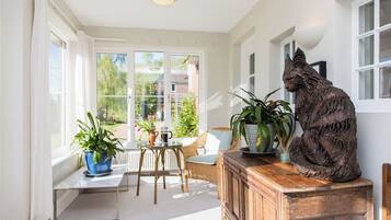 A sunlit room with large windows, a wicker chair, a wooden cabinet, and a cat statue.