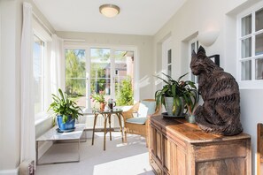 Una habitación iluminada por el sol, con amplios ventanales, una silla de mimbre, un aparador de madera y una estatua de un gato.