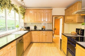 A kitchen with wooden cabinets, a black countertop, a dishwasher, a stove, and a window with a view of greenery. - Whitebeams (Canterbury)