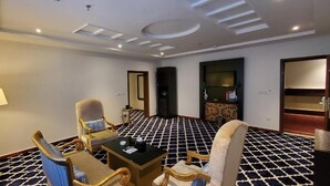 Habitación de hotel con una mesa de recepción, dos sillones, un sofá, una lámpara, un televisor y un suelo estampado.