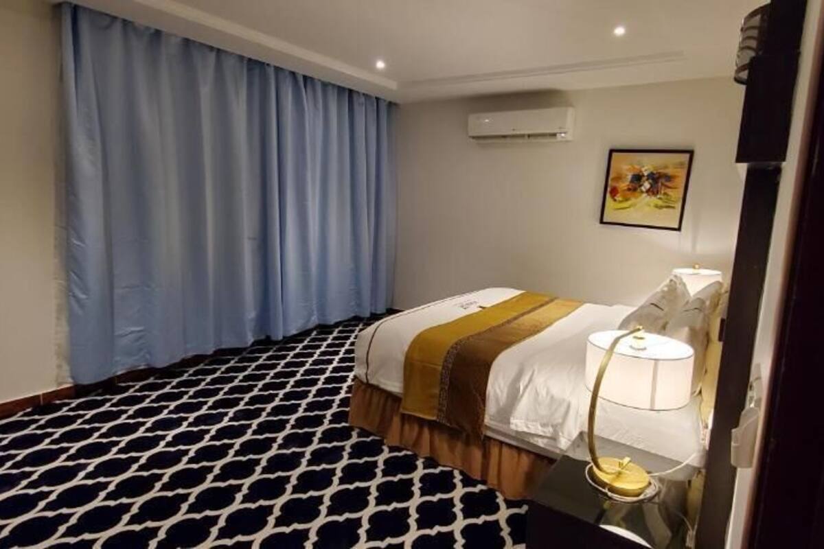 A hotel room with a patterned carpet, a bed with a white and brown bedspread, a bedside lamp, and a painting on the wall.