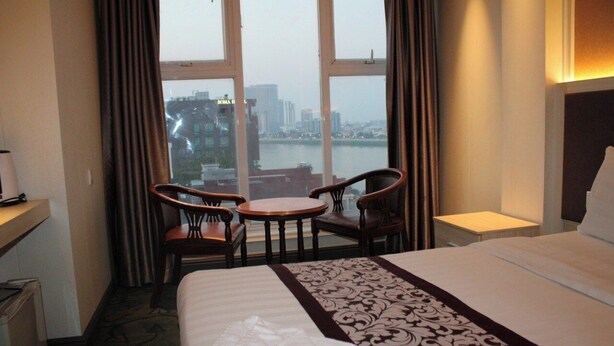 A hotel room with a bed, a desk, a chair, a small table, and a view of a cityscape through large windows. - Gold Indigo Hotel (Phnom Penh)
