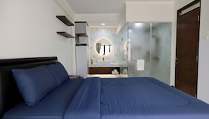 Una habitación de hotel moderna con una cama grande, un baño con cabina de ducha de vidrio y una pequeña zona de estar con un espejo redondo.