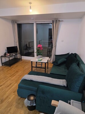A living room with a green sofa, a small table with a plant, a TV, and a sliding glass door leading to a balcony. - Berberis House (Feltham)