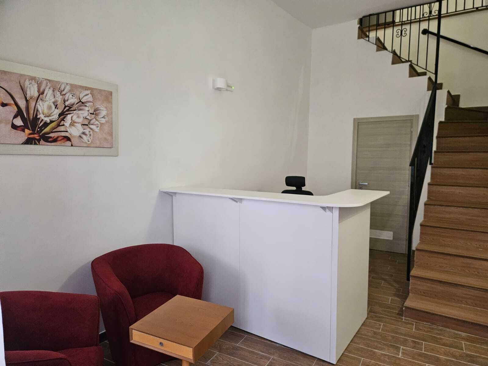 A room with a white desk, a red chair, a small table, a painting on the wall, and a staircase.