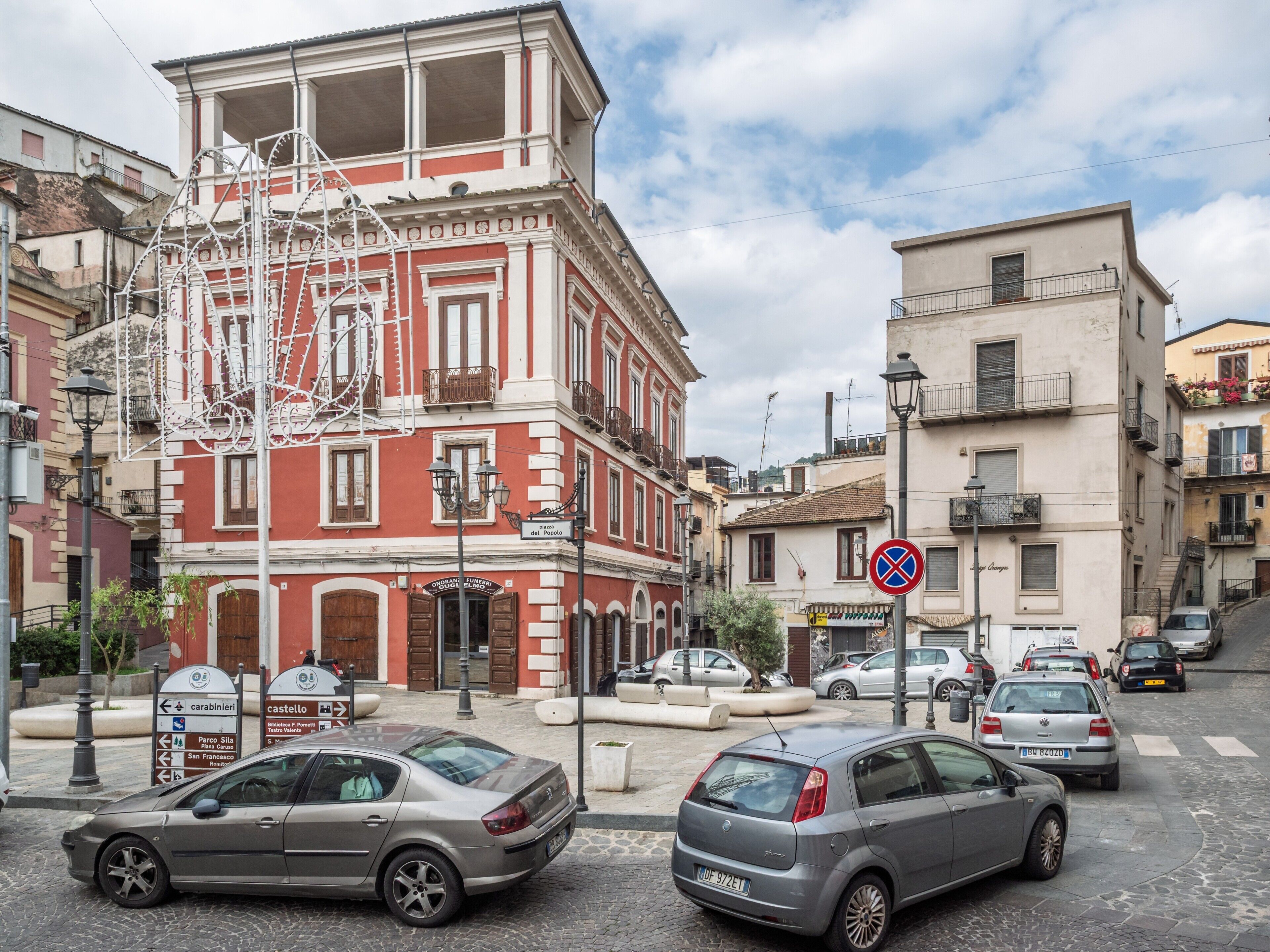 Una calle con coches aparcados, un edificio de fachada roja y un cartel de prohibido estacionar.