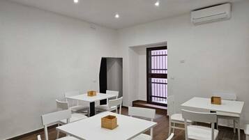 Una habitación con mesas y sillas blancas, un equipo de aire acondicionado y una ventana con barrotes.
