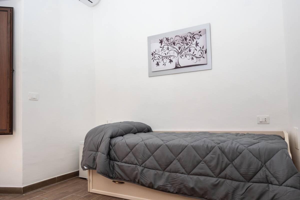 Un lit compact recouvert d’une housse matelassée, dans une pièce aux murs blancs et au plancher en bois.