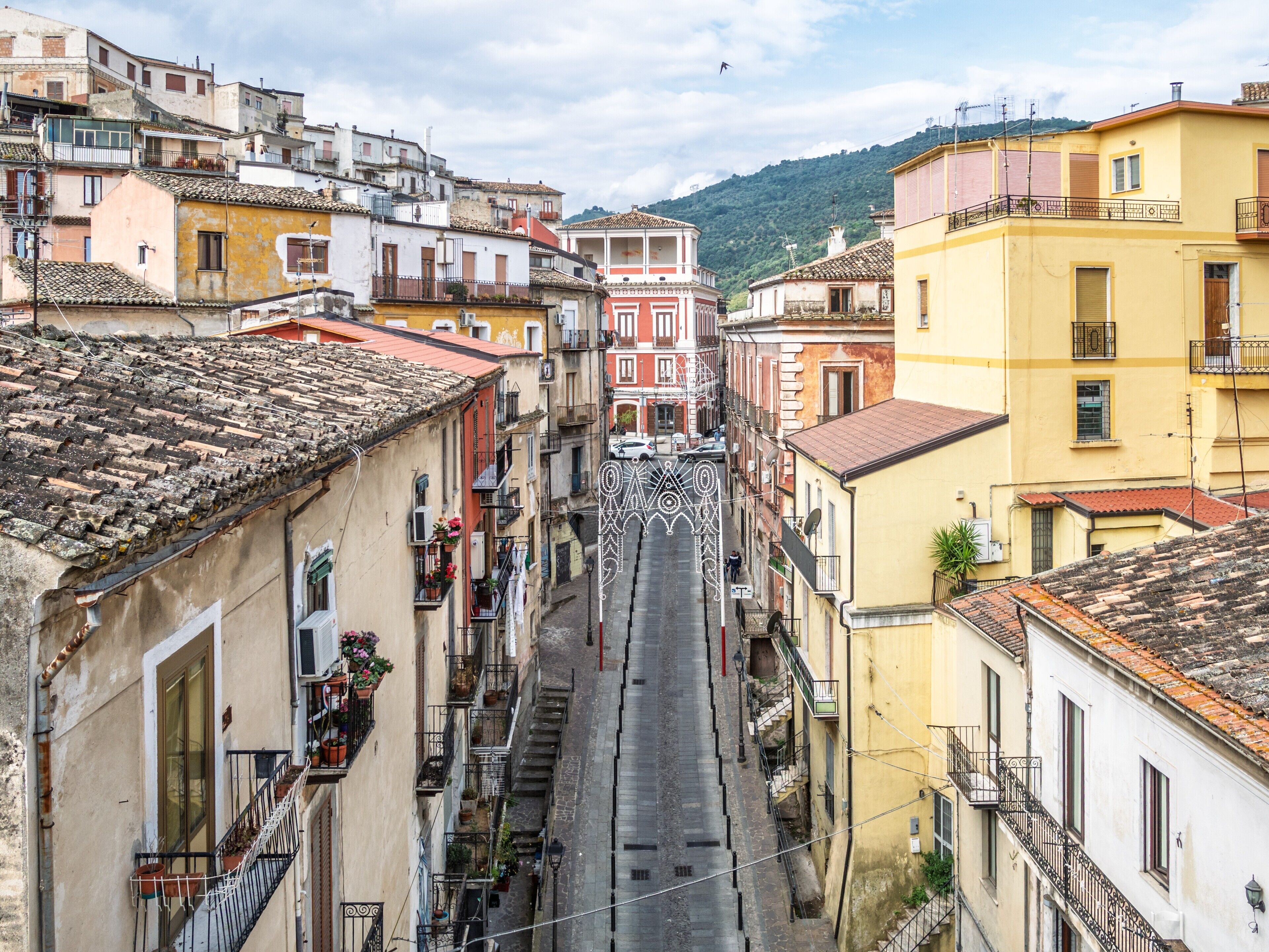 Una calle estrecha bordeada por edificios de varios pisos con balcones, un cielo despejado y un paisaje montañoso al fondo.