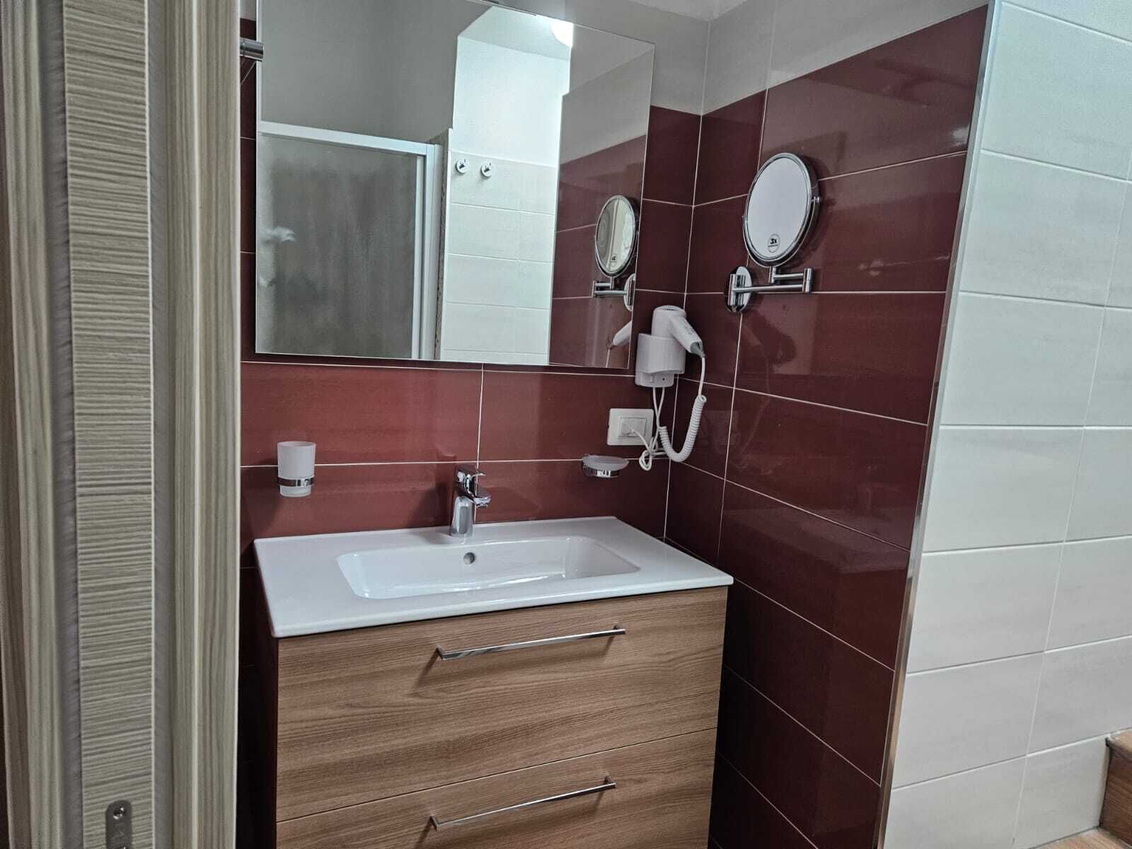 Ein Badezimmer mit weißem Waschbecken, Holzschrank, zwei runden Spiegeln, einer Handbrause und einem Handtuchhalter.