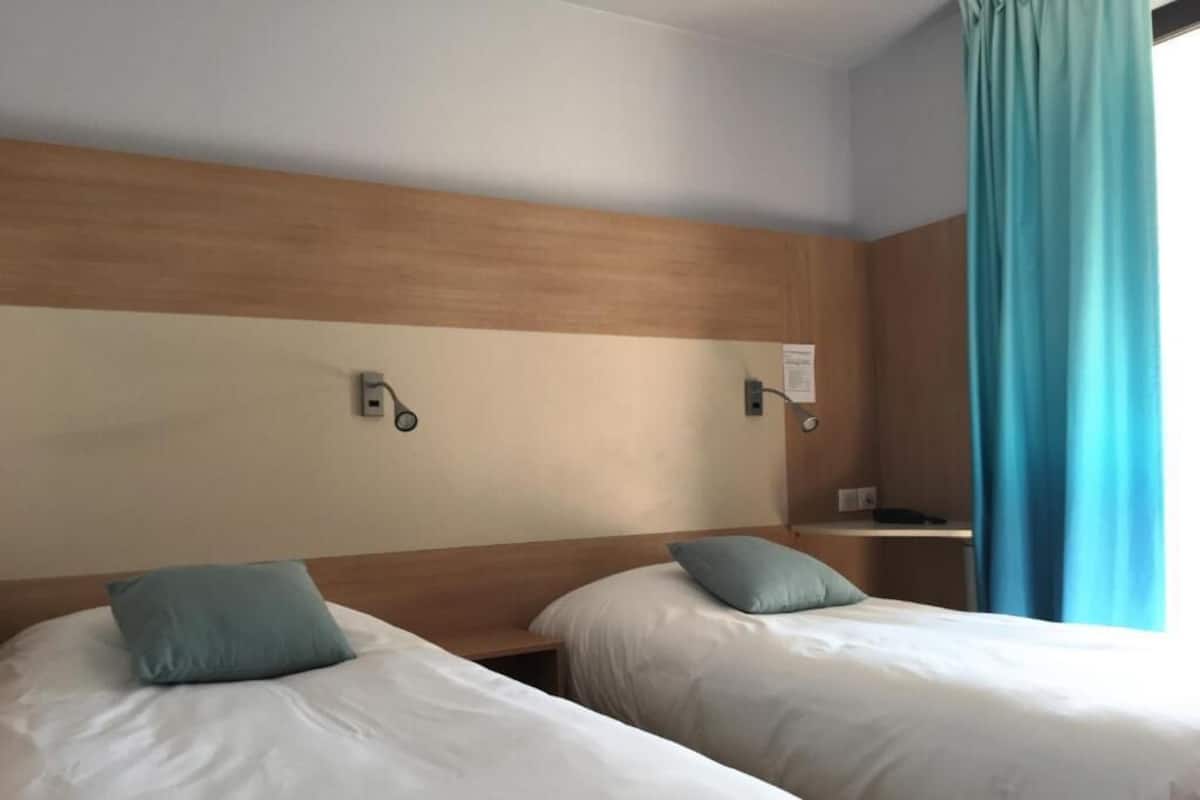 A hotel room with two beds, wooden headboard, a nightstand, and a window with a blue curtain.