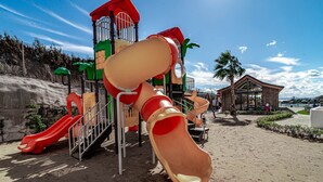 Ein Spielplatz mit Rutschen und Klettergerüsten, sandigem Untergrund und einem Gebäude mit geneigtem Dach im Hintergrund.