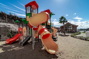 Ein Spielplatz mit Rutschen und Klettergerüsten, sandigem Untergrund und einem Gebäude mit geneigtem Dach im Hintergrund.