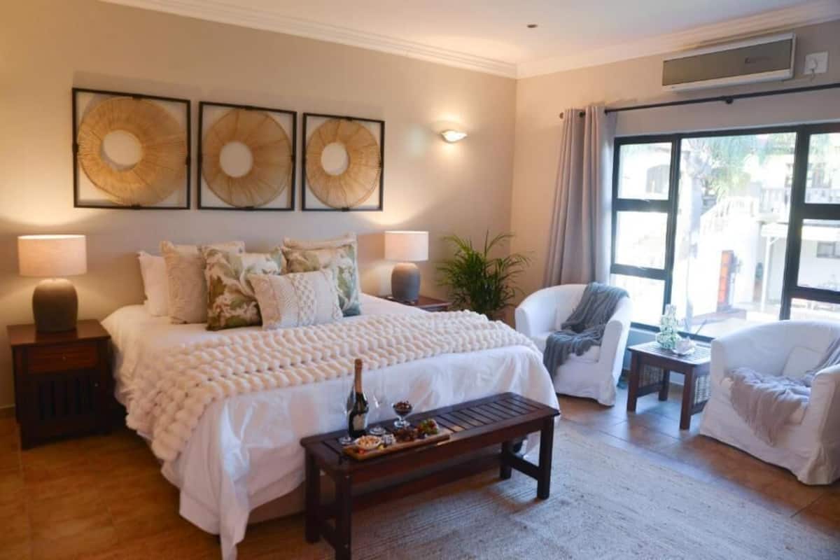 Un dormitorio con una cama grande, lámparas de noche, una mesita de noche, un banco de madera con aperitivos, una silla y una ventana con cortinas.