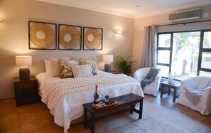 Un dormitorio con una cama grande, lámparas de noche, una mesita de noche, un banco de madera con aperitivos, una silla y una ventana con cortinas.