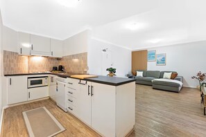Una cocina moderna con armarios blancos, un microondas y una cafetera. El suelo es de madera, hay un tapete pequeño y un sofá gris en la zona de estar.