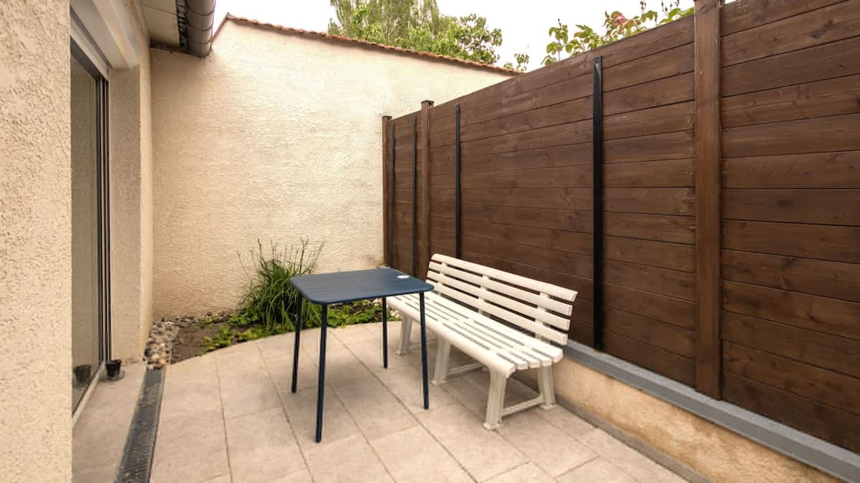 Un patio con una panchina bianca e un piccolo tavolo, una recinzione in legno e un edificio con facciata color crema.
