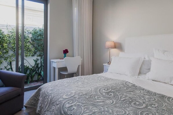 A hotel room with a bed, a desk, a chair, a lamp, and a view of greenery through a window. - 23 Grace (Cape Town)