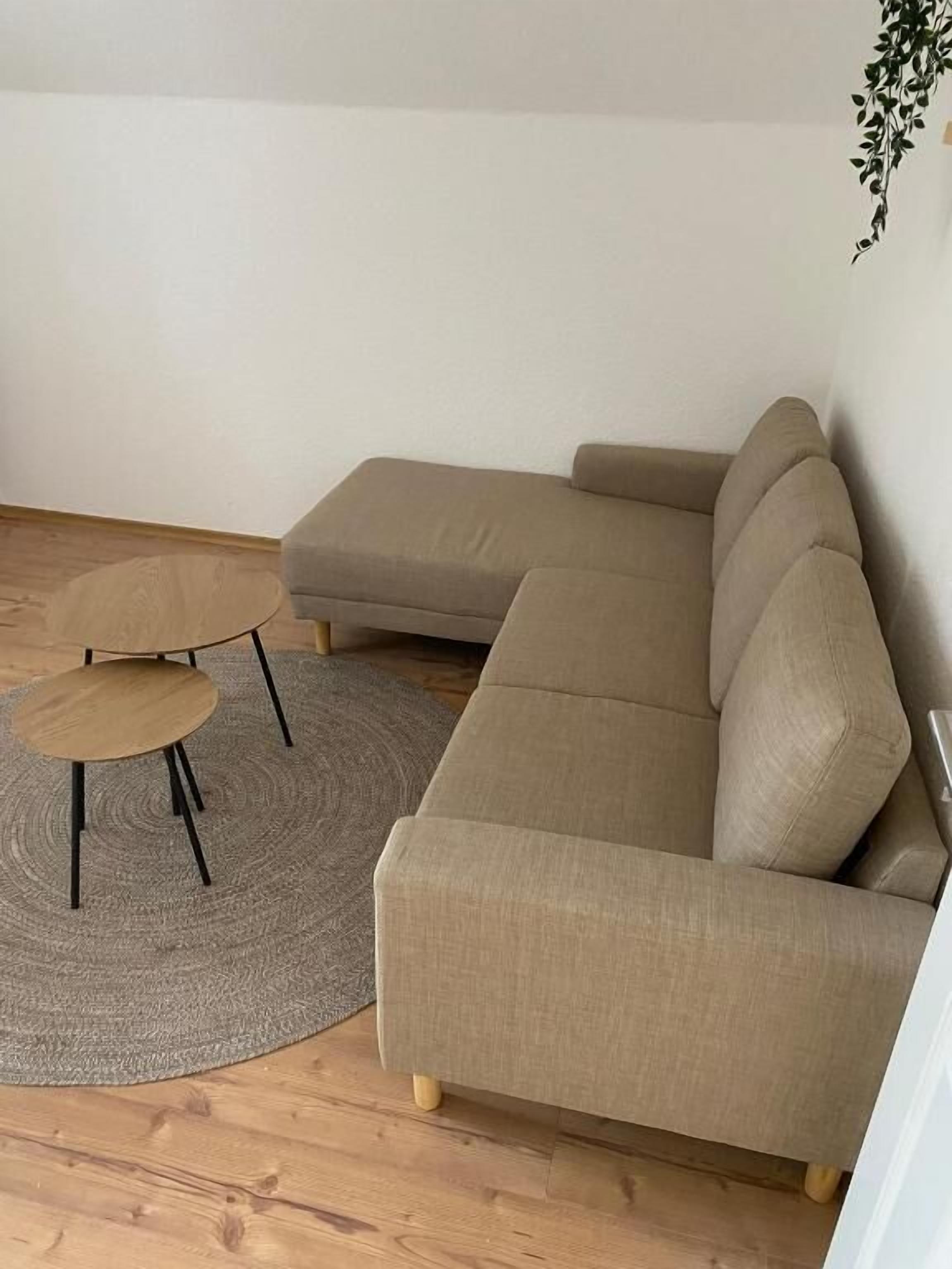 A beige sofa with a matching ottoman, a small round table, and a woven rug in a room with wooden flooring and a white wall.