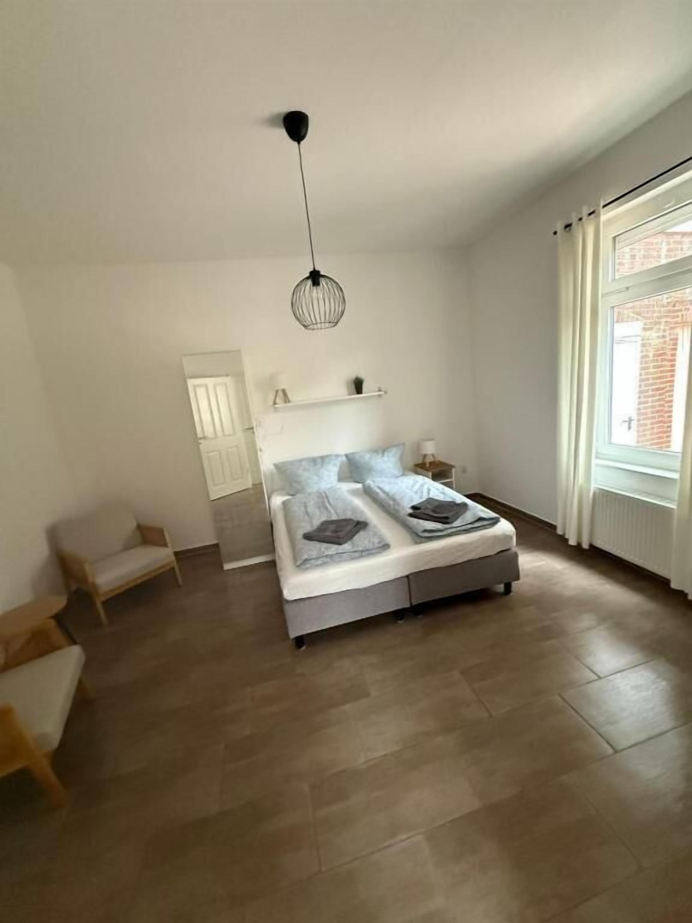 Un dormitorio con cama, una lámpara colgante, una ventana con cortinas, una puerta y una mesita.
