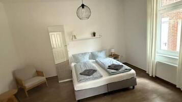 Un dormitorio con cama, una lámpara colgante, una ventana con cortinas, una puerta y una mesita.
