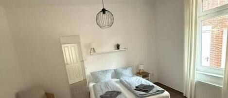 A bedroom with a bed, a hanging light fixture, a window with curtains, a door, and a small table.