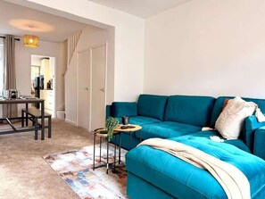 Ein modernes Wohnzimmer mit einem blaugrünen Sofa, einem kleinen runden Tisch mit einer Pflanze und einem farbenfrohen Teppich.
