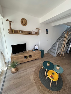 Un salon comprenant un meuble de rangement en bois pour l’équipement audiovisuel, un téléviseur, une petite table avec des chaises, un escalier et une étagère murale agrémentée d’objets décoratifs.