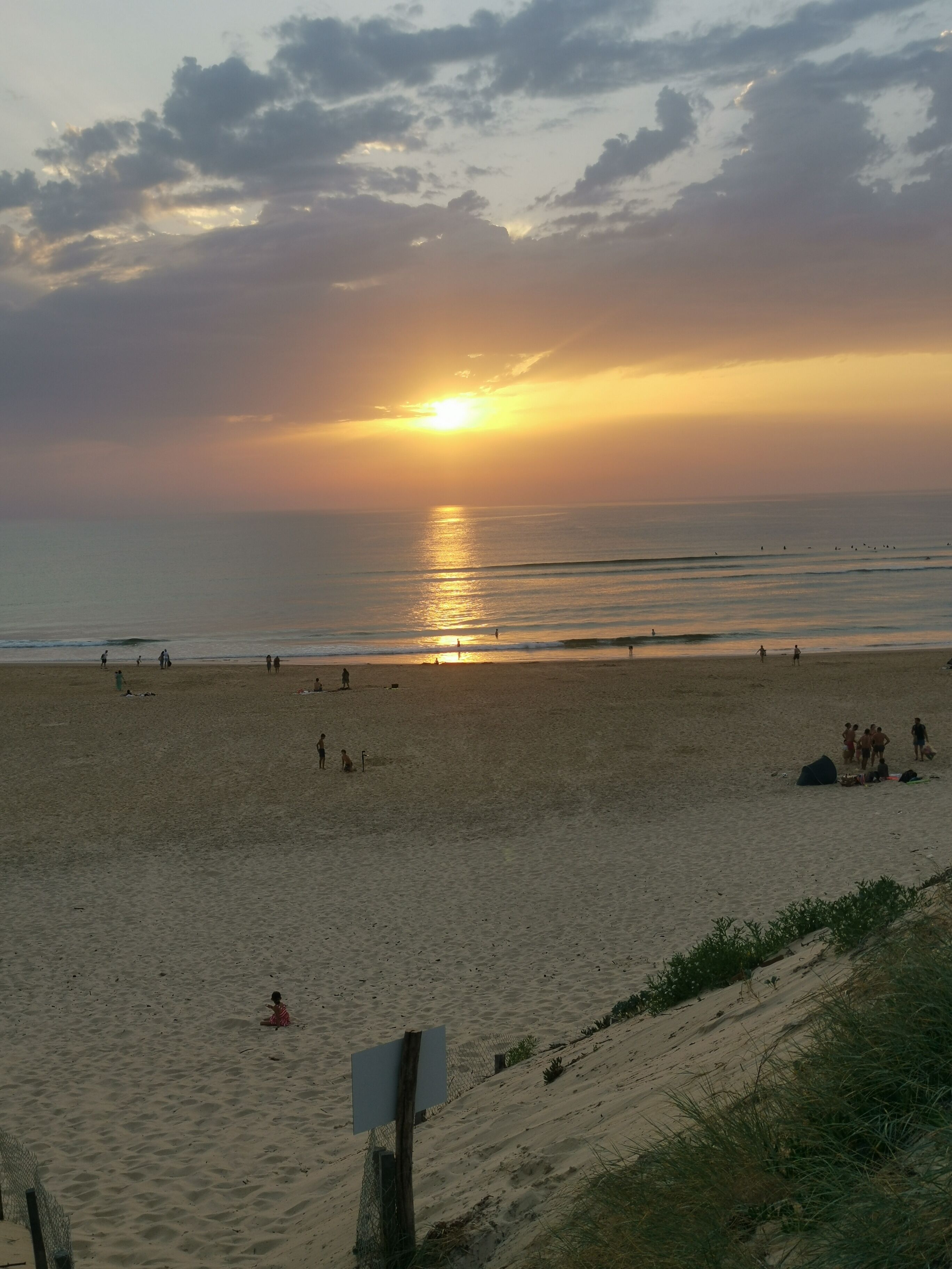 Een strand bij zonsondergang, mensen wandelen en een bord.