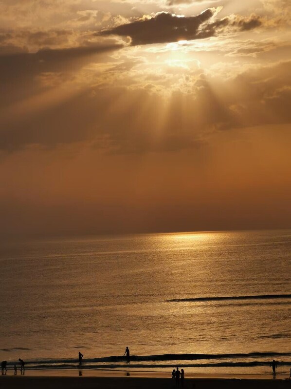 A beach scene with the sun setting over the ocean, silhouettes of people on the shore, and a calm sea.