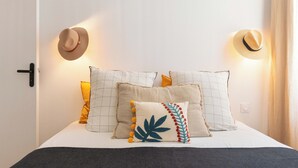 Uma cama com roupa de cama branca, várias almofadas e dois chapéus fixados na parede.
