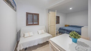 Ein kleiner, hell eingerichteter Raum mit einem Bett, einem weißen Sofa, einem Tisch und einer Topfpflanze.