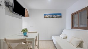 Ein Zimmer mit einer weißen Couch, einem Esstisch mit Stühlen, einem an der Wand befestigten Fernseher und einem Bild.