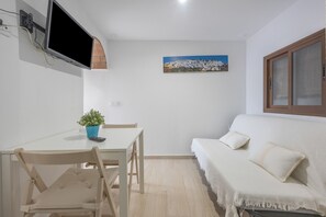 Een kamer met een witte bank, een eettafel met stoelen, een aan de muur bevestigde televisie en een schilderij.