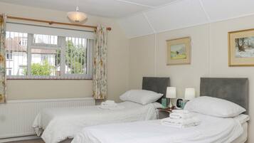 Une chambre d’hôtel avec deux lits, un radiateur, une fenêtre avec des rideaux, un luminaire au plafond et deux tableaux encadrés accrochés au mur.