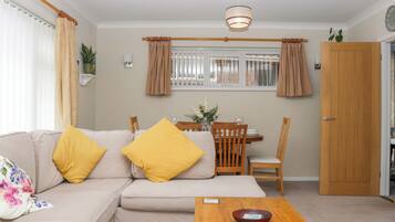 A living room with a sofa, a wooden coffee table, a dining table with chairs, a window with curtains, and a door.