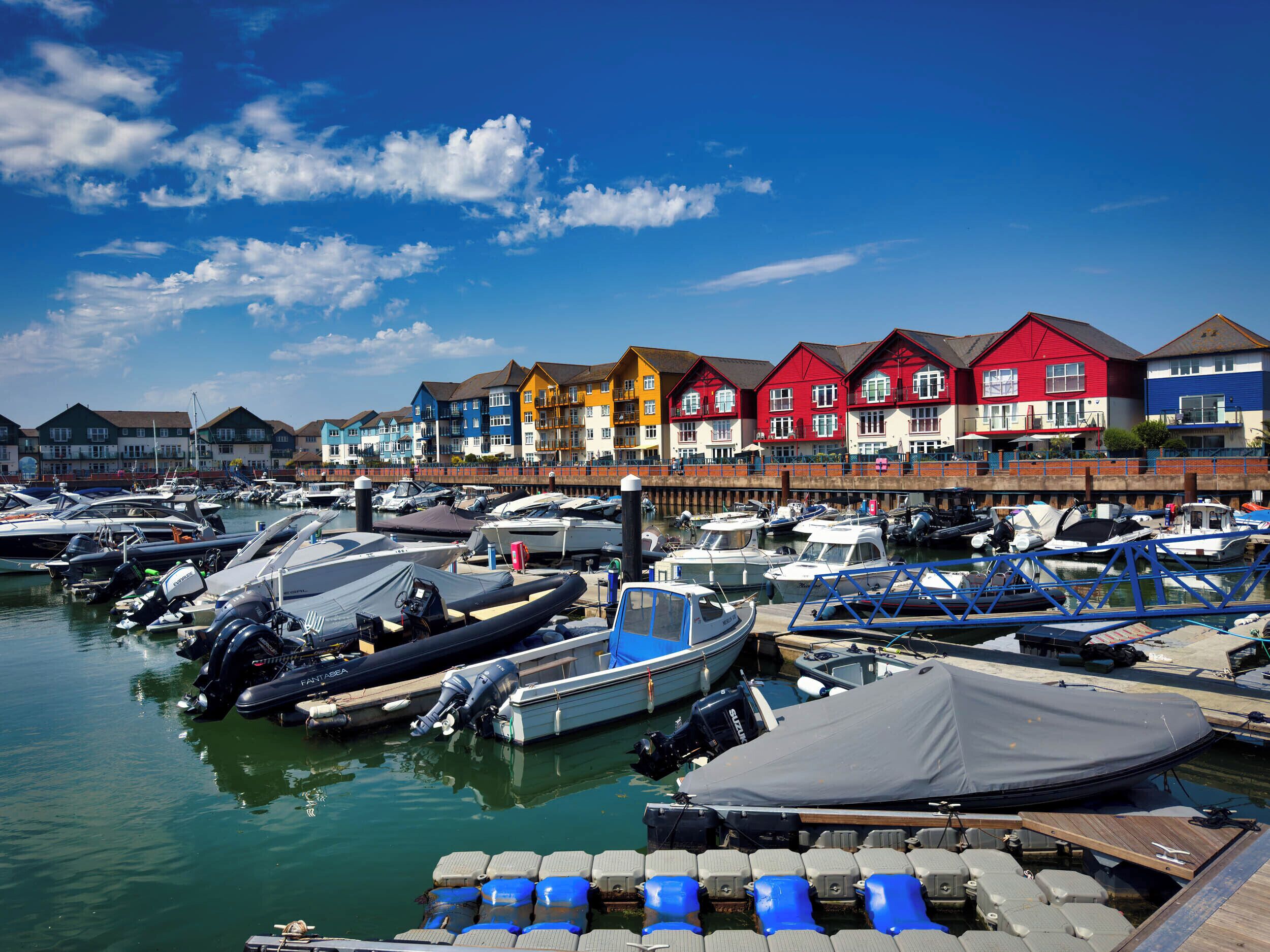 Un port esportiu amb vaixells amarrats, una filera de cases de colors i un cel blau i clar.