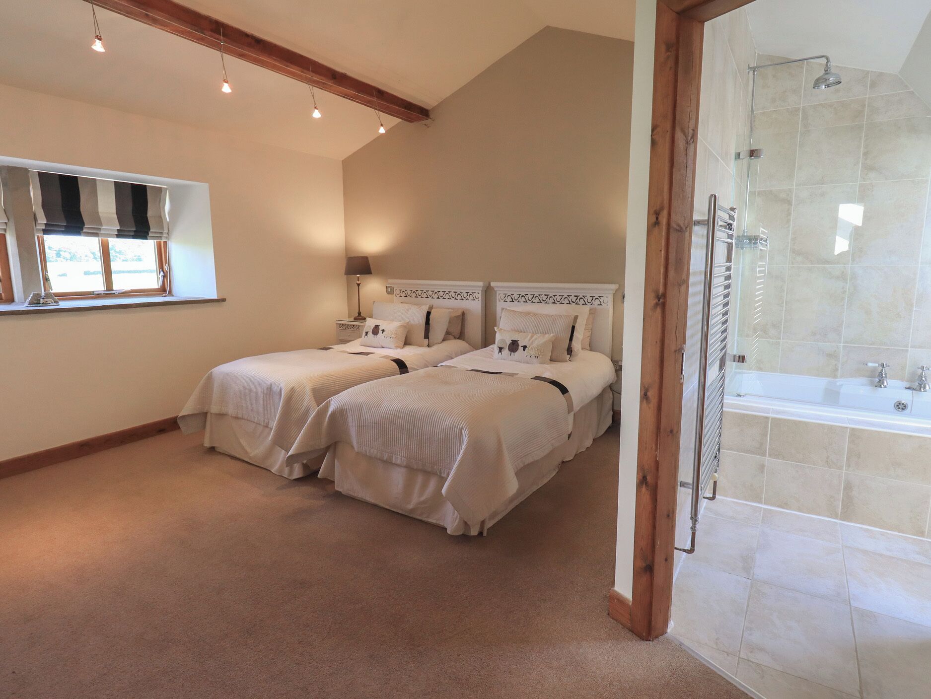 A bedroom with two beds, a window with blinds, a shower area with a glass door, and a wooden ceiling beam.