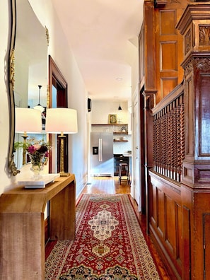 A hallway with a wooden floor, a red patterned rug, a wooden bench, and a mirror with a vase of flowers.