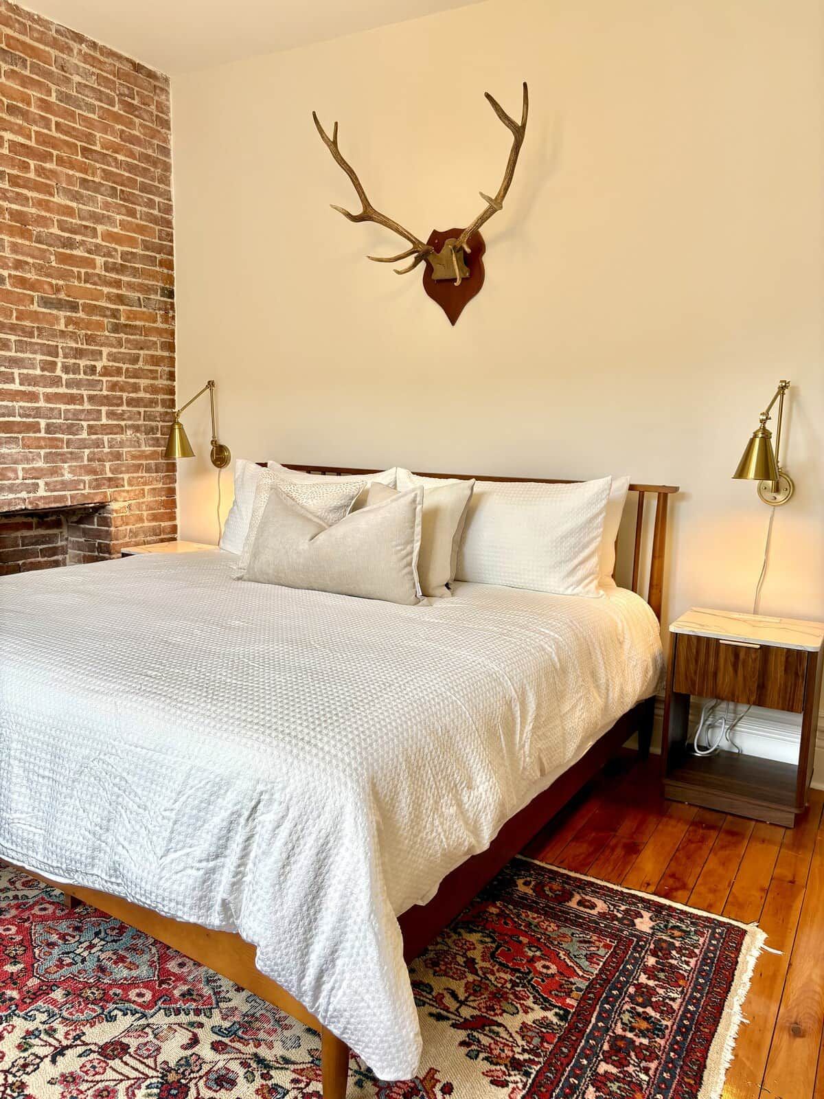 A bedroom with a brick wall, a bed with white bedding, a decorative antler headpiece on the wall, and a patterned rug on the floor.