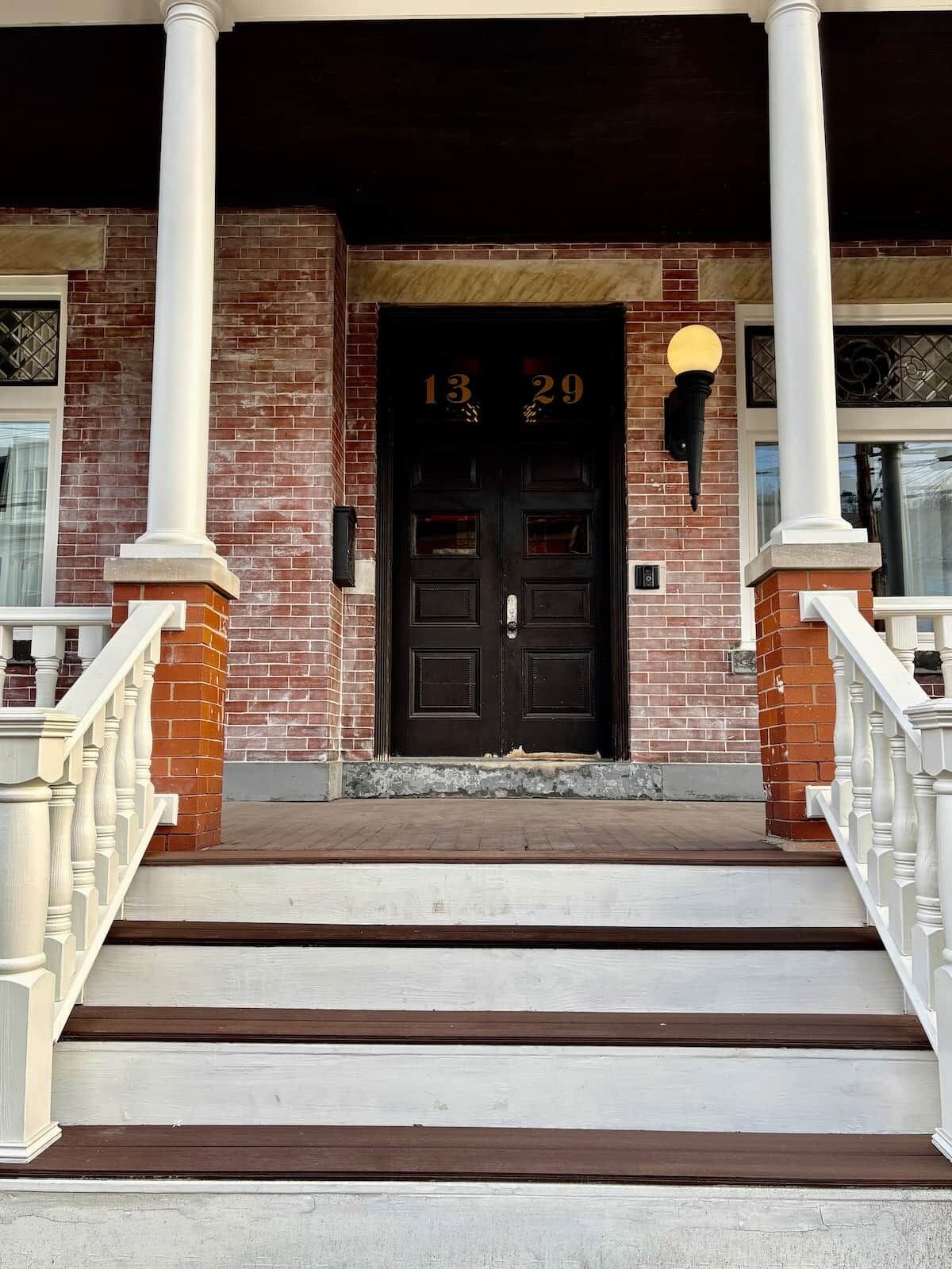 A brick building with a black double door, white columns, and a white railing on the steps leading up to the entrance.