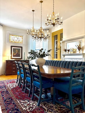 A dining room with a large wooden table surrounded by blue upholstered chairs, a chandelier, a large vase with greenery, and a patterned rug. - Middle Manor | Sleeps 8, with parking | 5mi to Pgh (Pittsburgh)