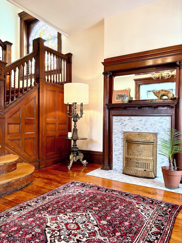 A grand staircase with ornate woodwork, a fireplace with a decorative mantel, and a patterned rug on the floor.