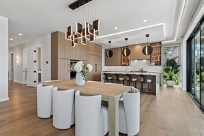 A modern kitchen with a central island, pendant lighting, and a dining area with a wooden table and white chairs. - Luxury waterfront home: dock, pool, spa, golf sim (Indian Rocks Beach)