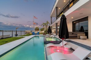 A modern outdoor pool area with lounge chairs, a hot tub, and a view of the ocean. - Luxury waterfront home: dock, pool, spa, golf sim (Indian Rocks Beach)