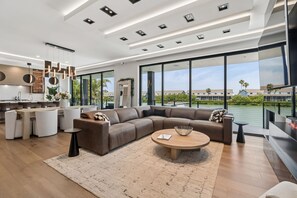 A modern living room with a sectional sofa, a round coffee table, and a large window offering a view of the water and buildings. - Luxury waterfront home: dock, pool, spa, golf sim (Indian Rocks Beach)