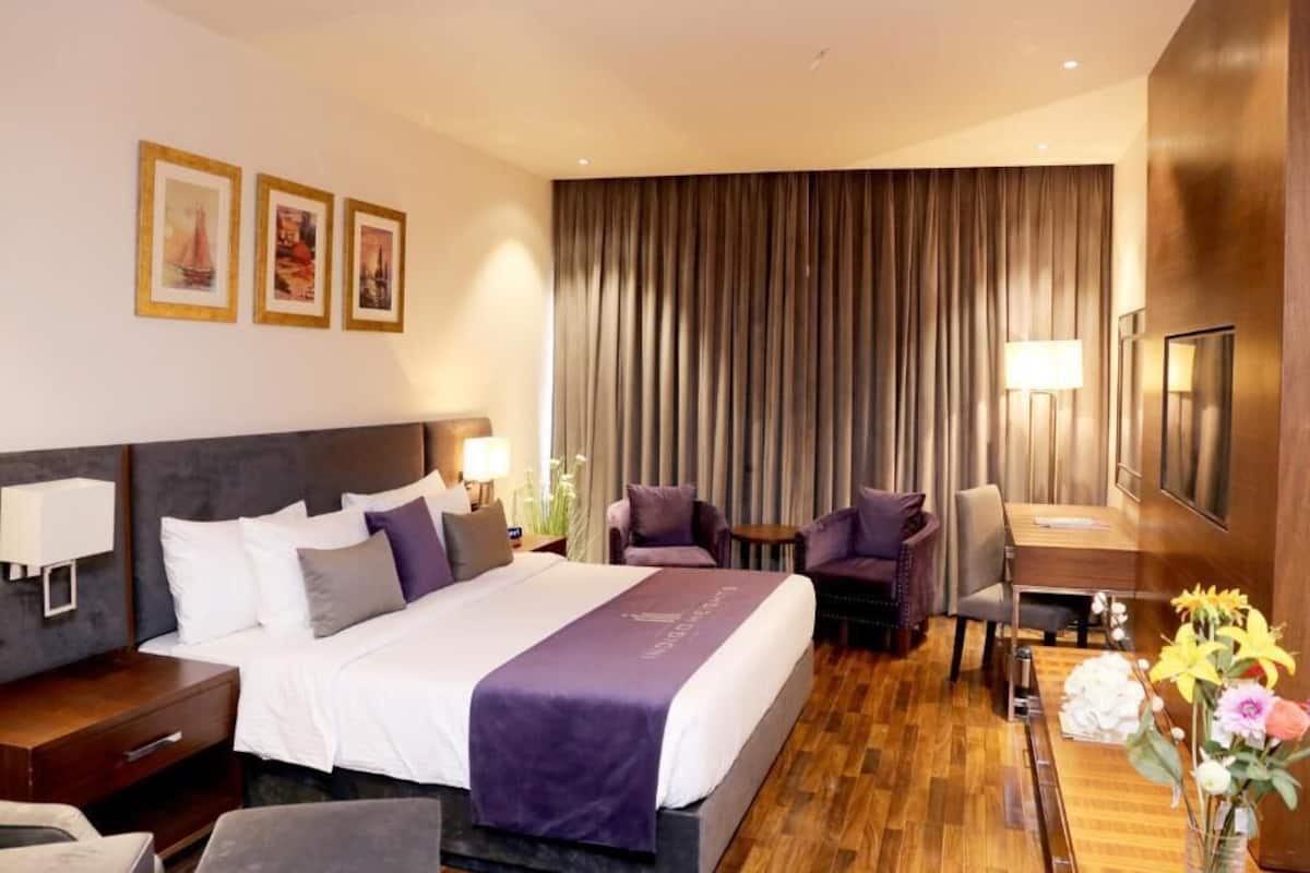 A hotel room with a large bed, two armchairs, a small table, a desk, and a TV.