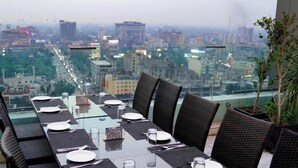 A dining area with a city view, glass table, wicker chairs, and a potted plant. - Indigo Heights Hotel and Suites (Lahore)