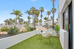 Un balcon avec un revêtement de gazon synthétique, un mur blanc, une chaise en osier et une vue sur des palmiers et des immeubles.